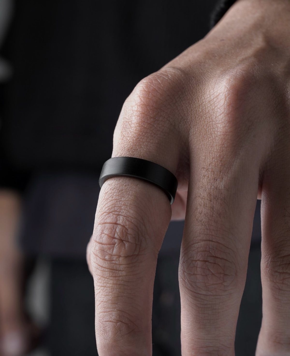 A matte black ring worn on a man's finger against a dark background.