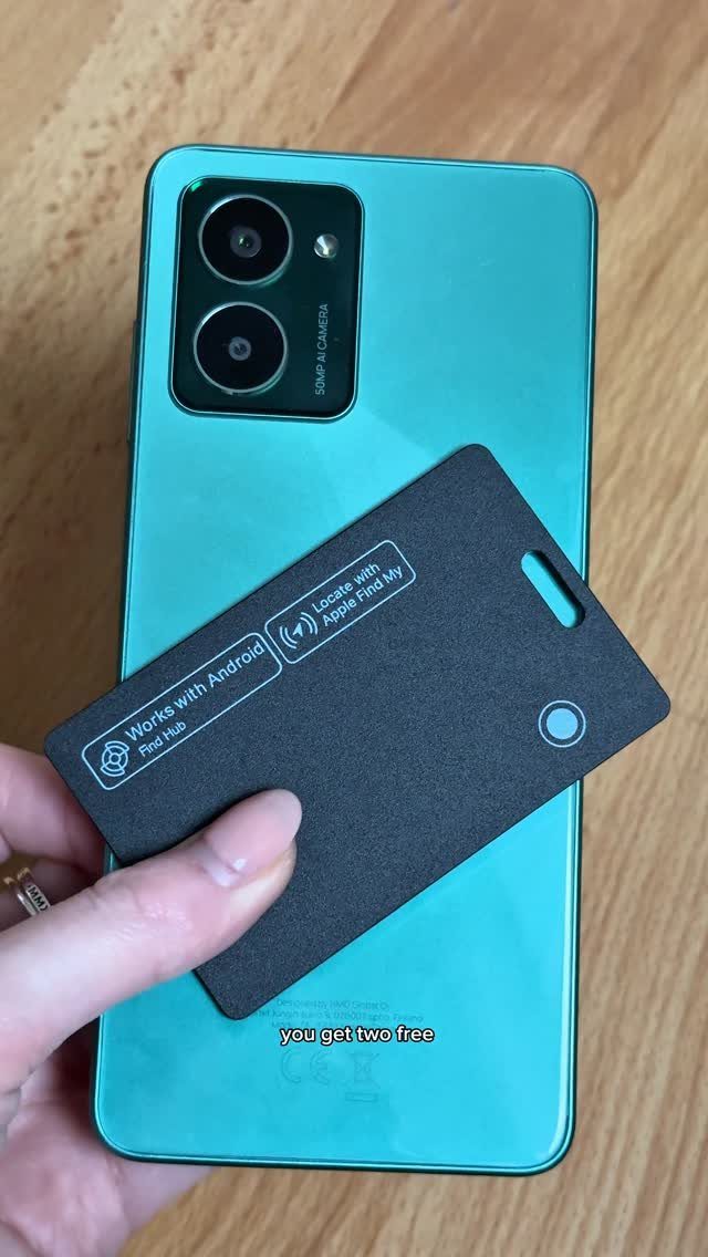 A hand holding a black tracker card over a light blue smartphone on wood.