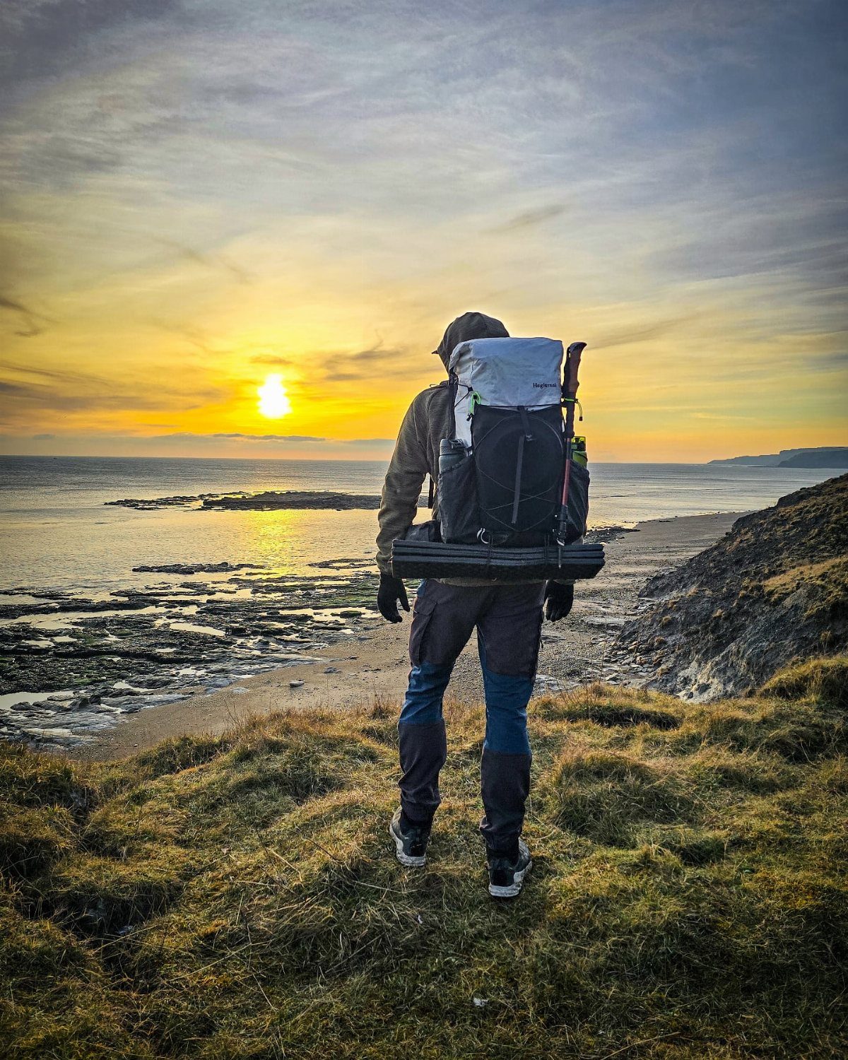 A hiker with a large backpack standing on a grassy cliff overlooking the ocean at sunset.