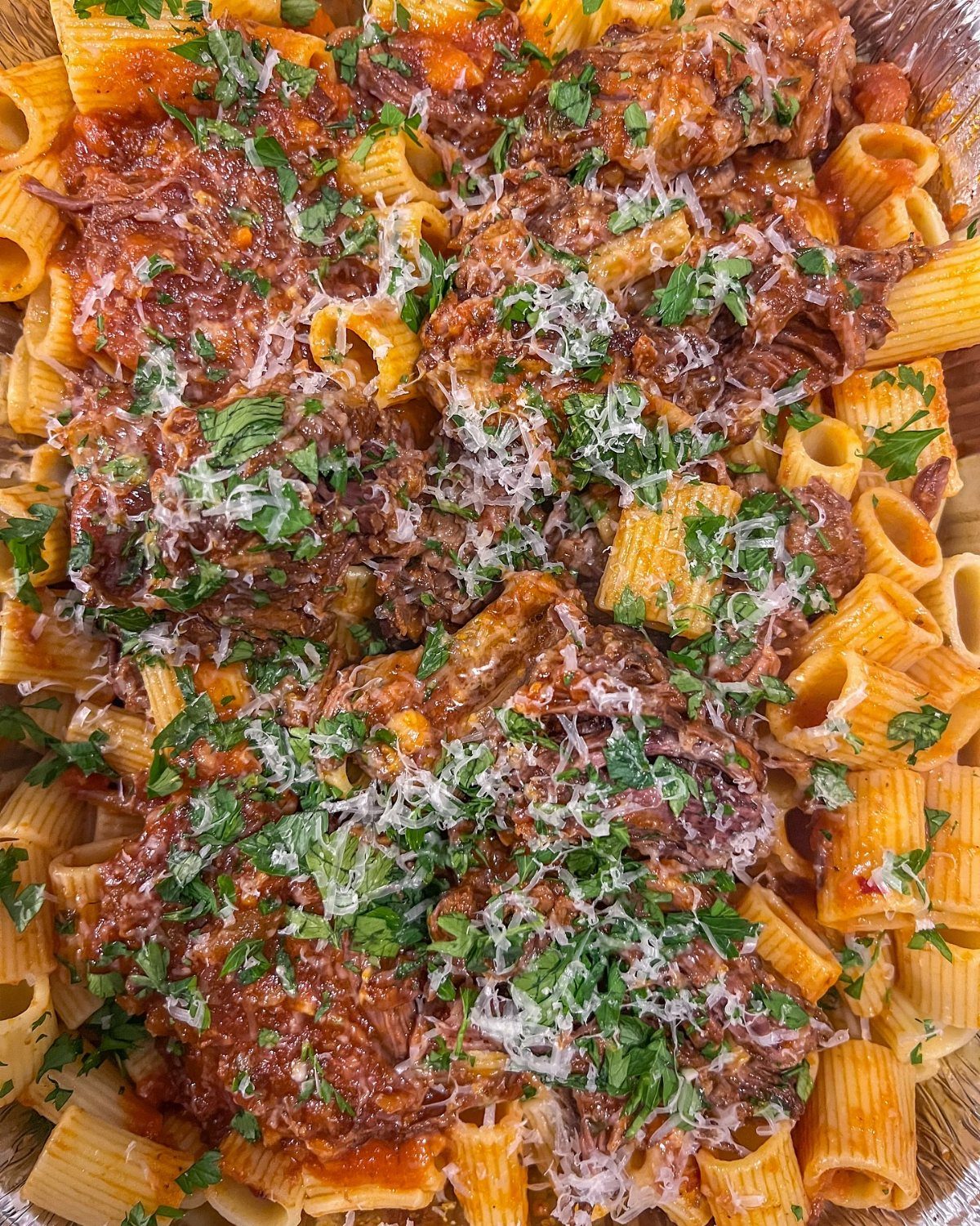 Rigatoni pasta topped with meat ragu, grated cheese, and fresh herbs in a metal dish.