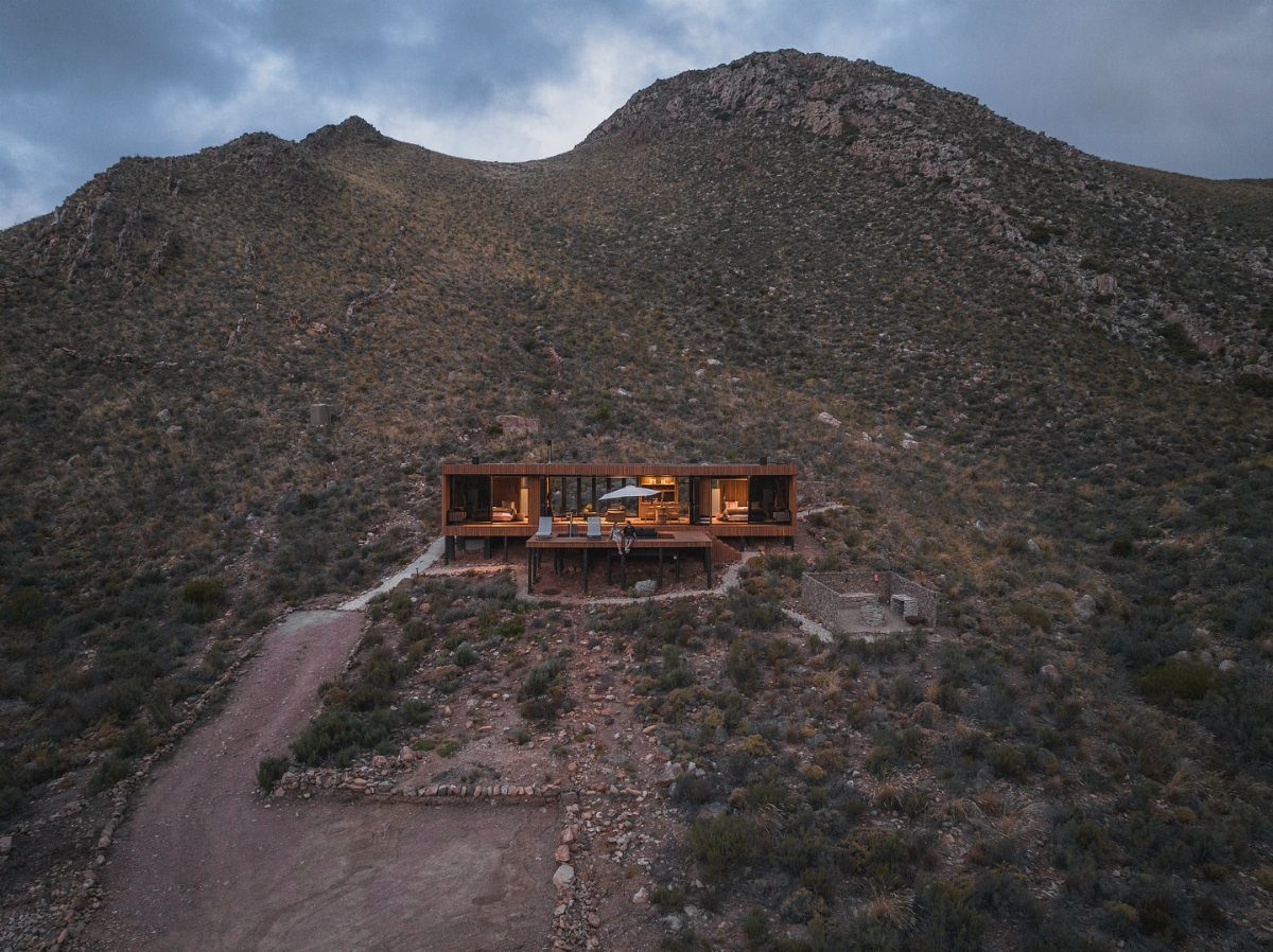 A modern wooden cabin with glowing windows nestled at the base of a large mountain.