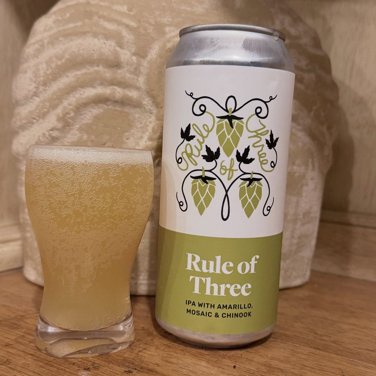 A can of Rule of Three IPA next to a glass of hazy yellow beer on wood.