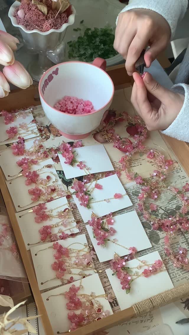 Hands assembling pink floral jewelry next to a mug of pink beads on a tray.
