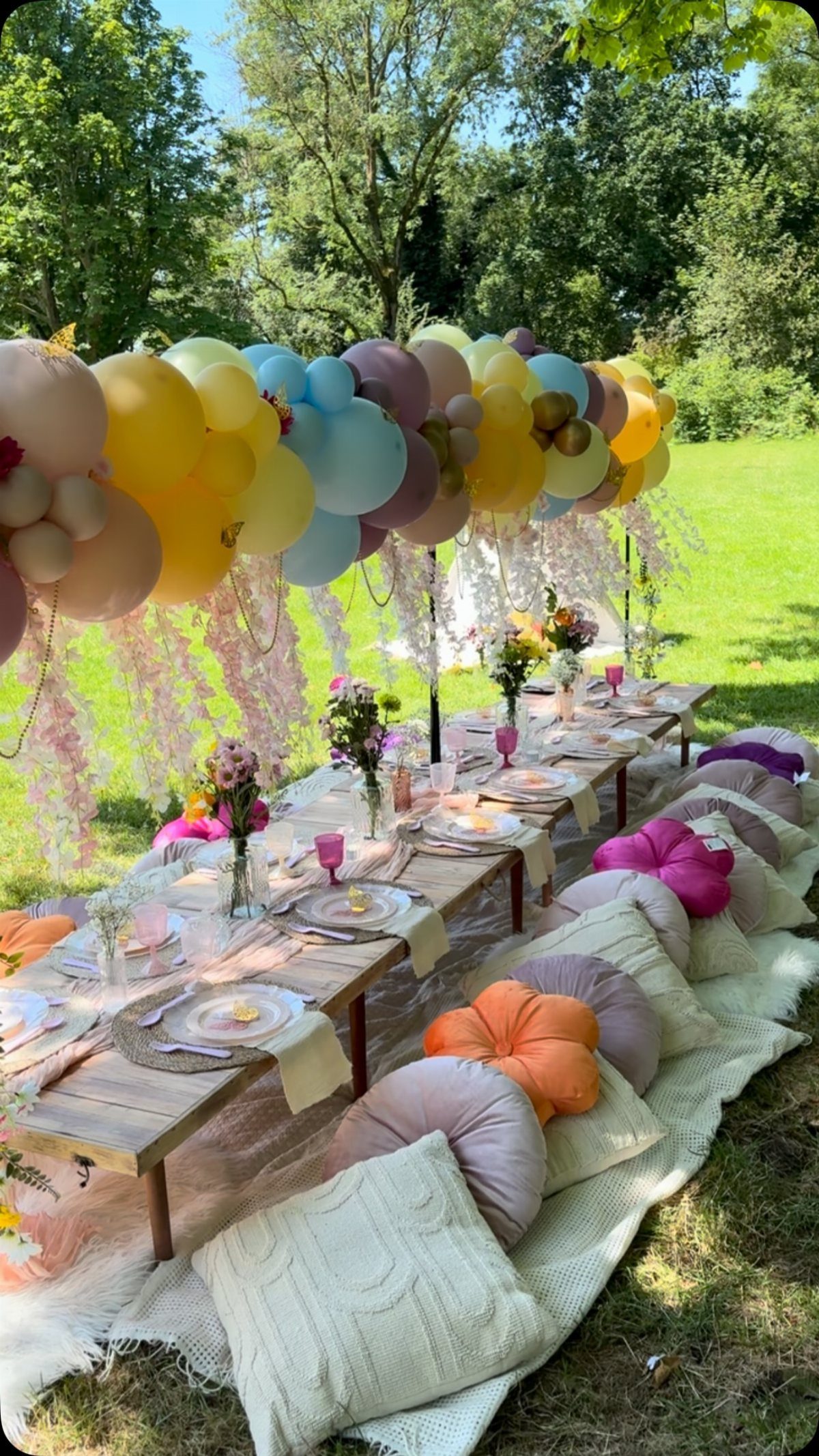 A decorated outdoor dining table with colorful balloons and floor pillows on a green lawn.