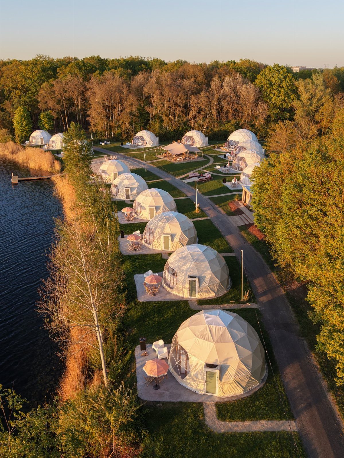 Geodesic dome tents arranged along a lakeside path surrounded by green trees.