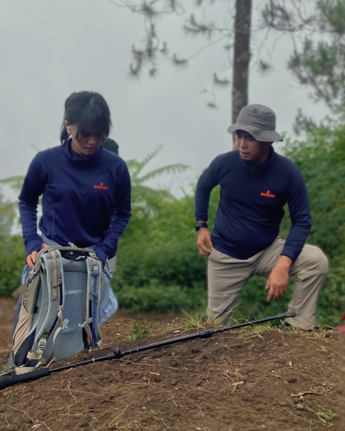 Two people preparing hiking gear with a backpack and trekking pole in a misty forest.