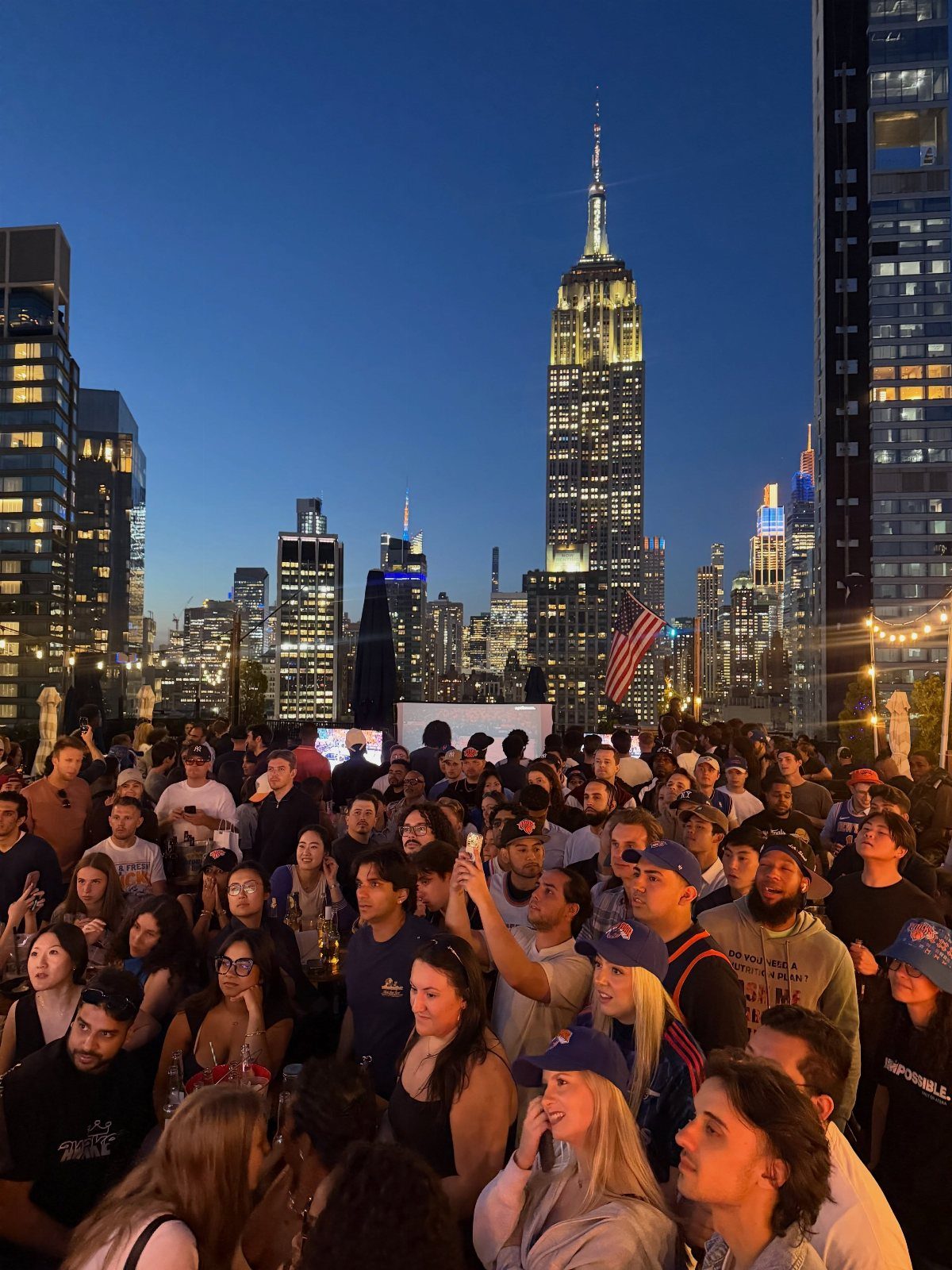 A crowd of people gathered on a rooftop overlooking a lit city skyline at dusk.