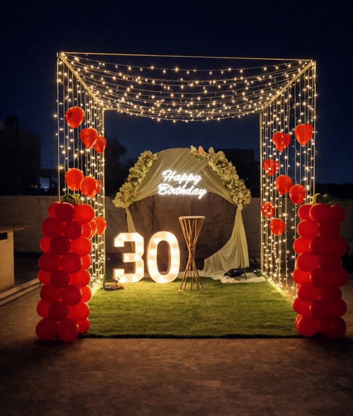 Illuminated 30 birthday decorations with red balloons, fairy lights, and a neon sign on grass.