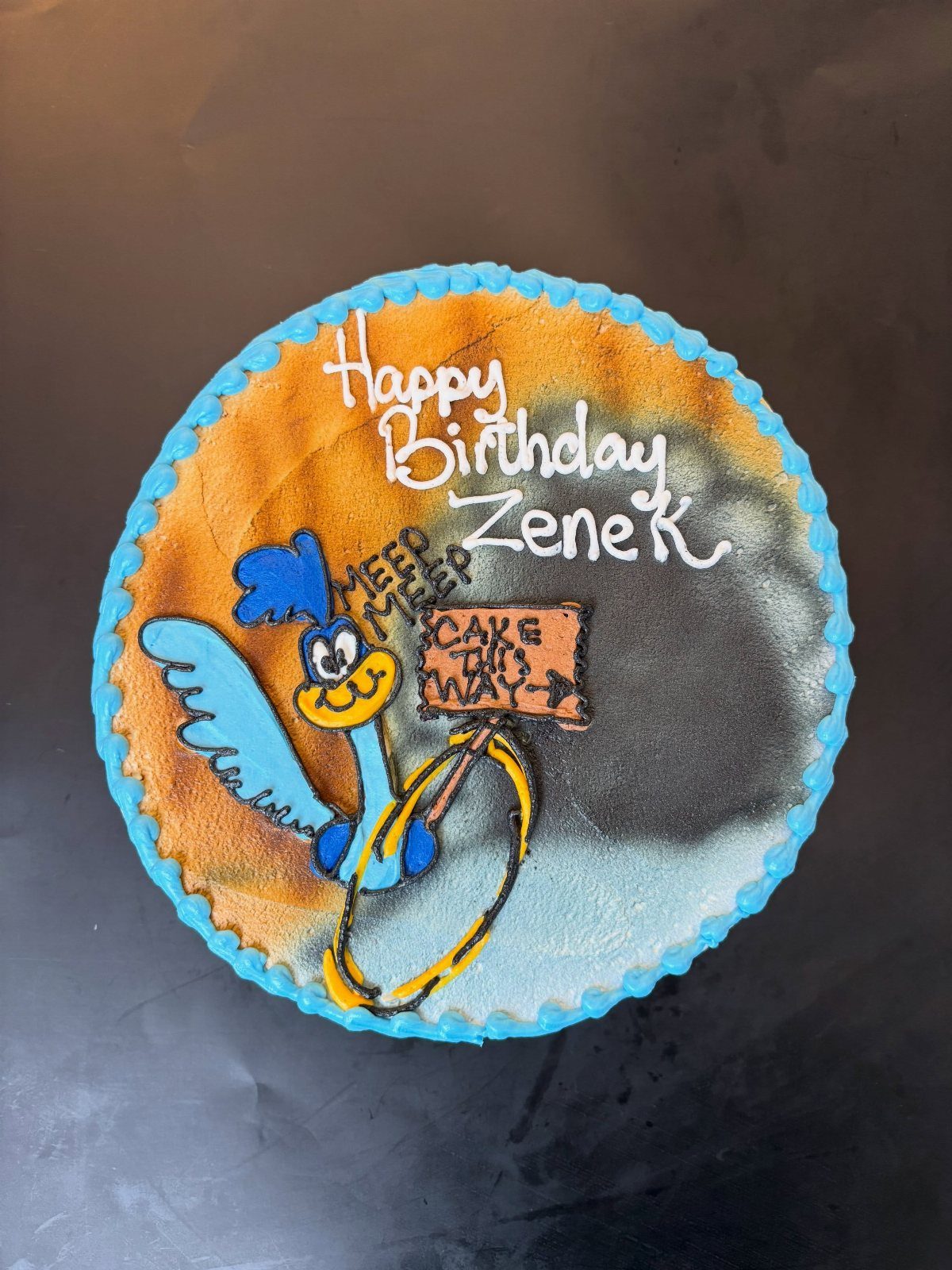 A round birthday cake featuring a blue cartoon bird character on an orange and grey background.