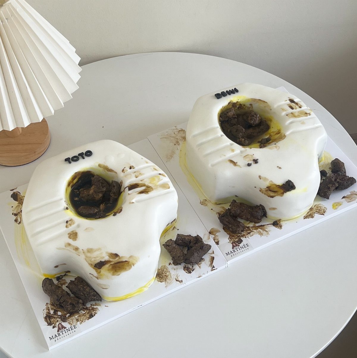 Two white toilet-shaped novelty cakes with dark edible decorations on a white table.