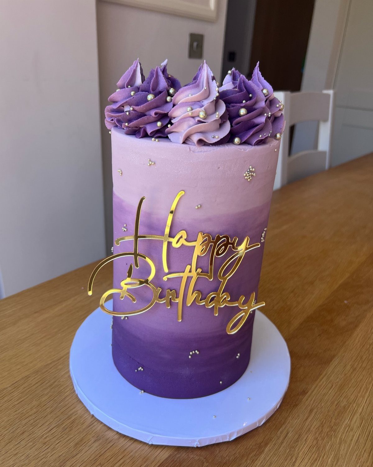 Purple ombre birthday cake with gold lettering and decorative frosting swirls on a wooden table.