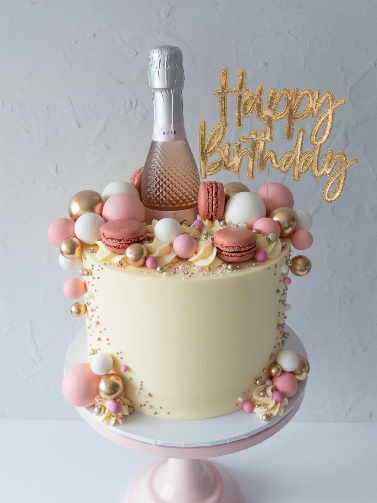 Cream cake decorated with pink macarons, gold spheres, a mini champagne bottle, and a gold topper.