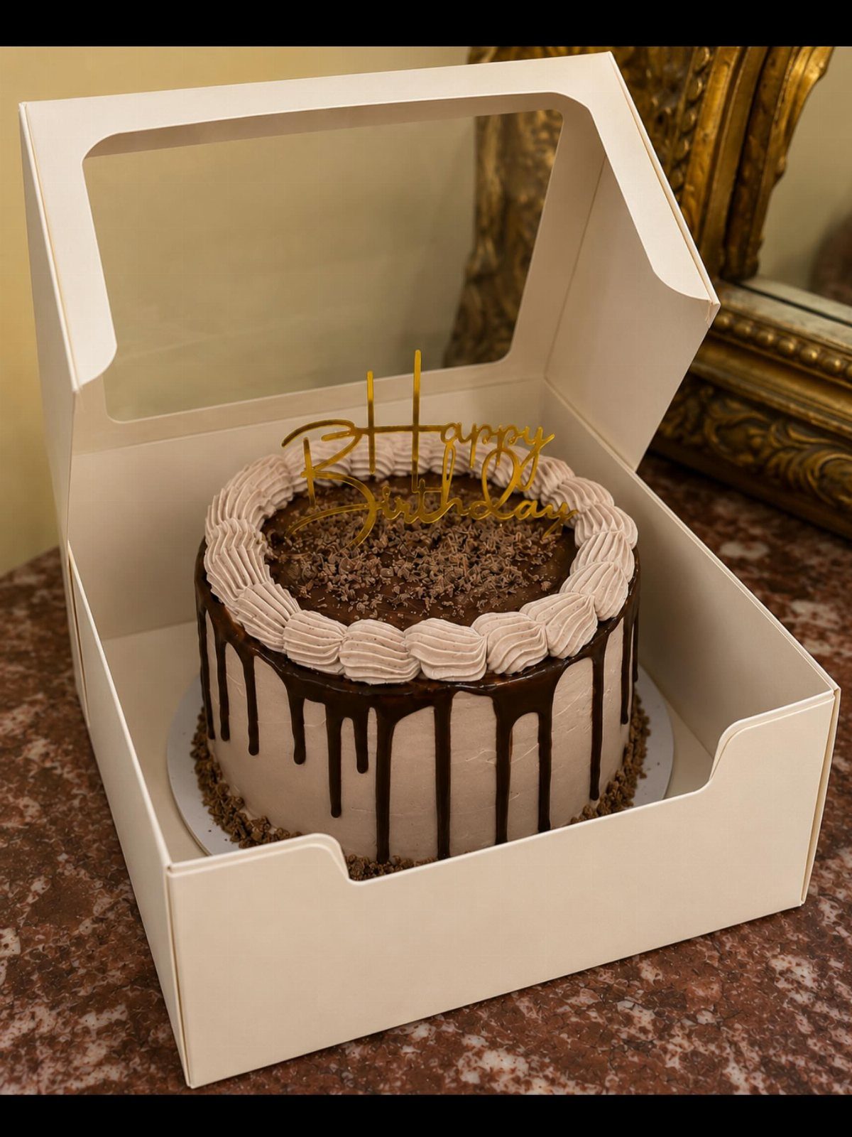 Chocolate drip cake with gold birthday topper inside an open white box.