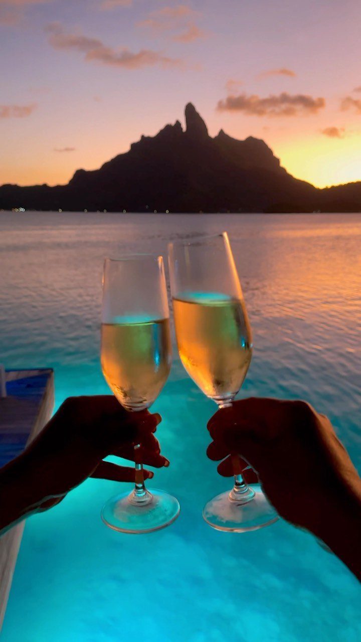 Two hands toast with champagne flutes against a sunset mountain backdrop over calm water.