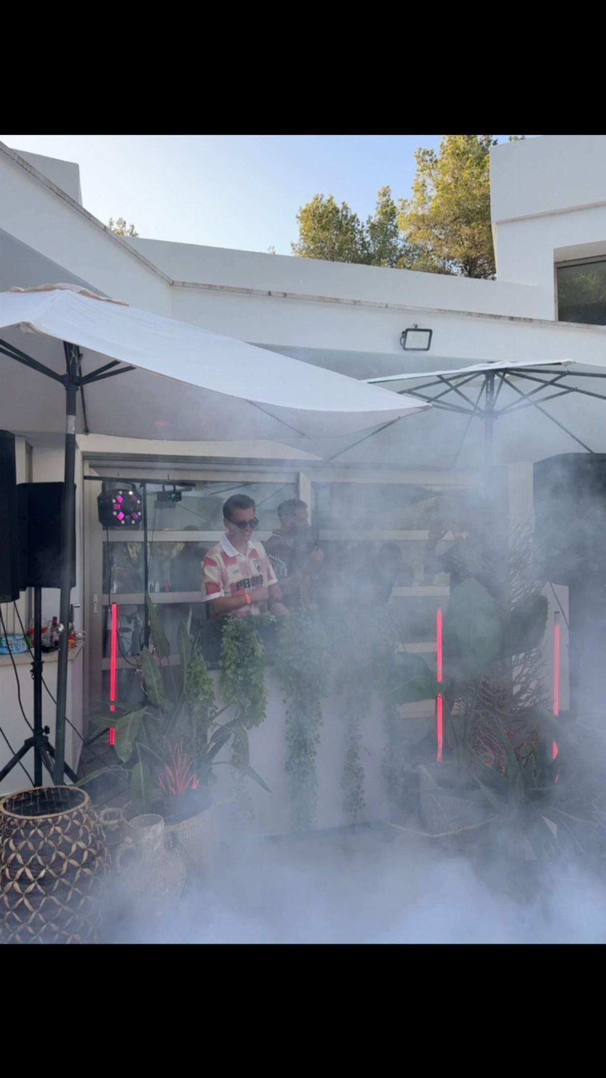 A DJ performs behind equipment amidst thick white smoke at an outdoor party.