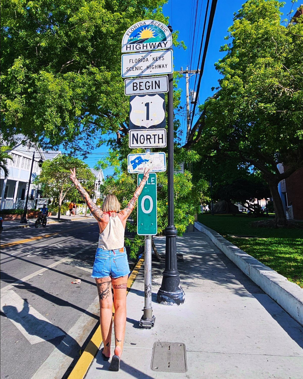 Woman with tattoos standing with arms raised facing the Florida Keys Mile 0 sign.
