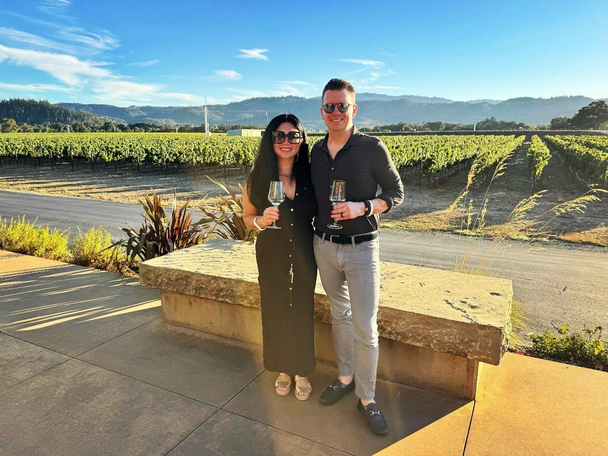 A man and woman holding wine glasses while posing in front of a sunny Napa Valley vineyard.