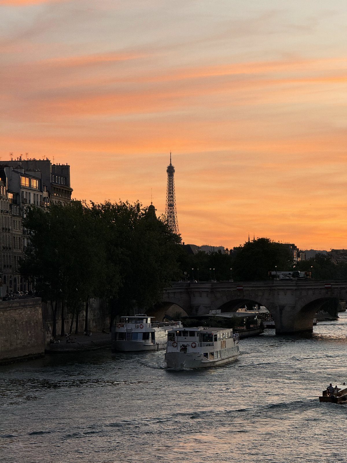 Eiffel Tower silhouetted against a colorful sunset over the Seine River with boats.