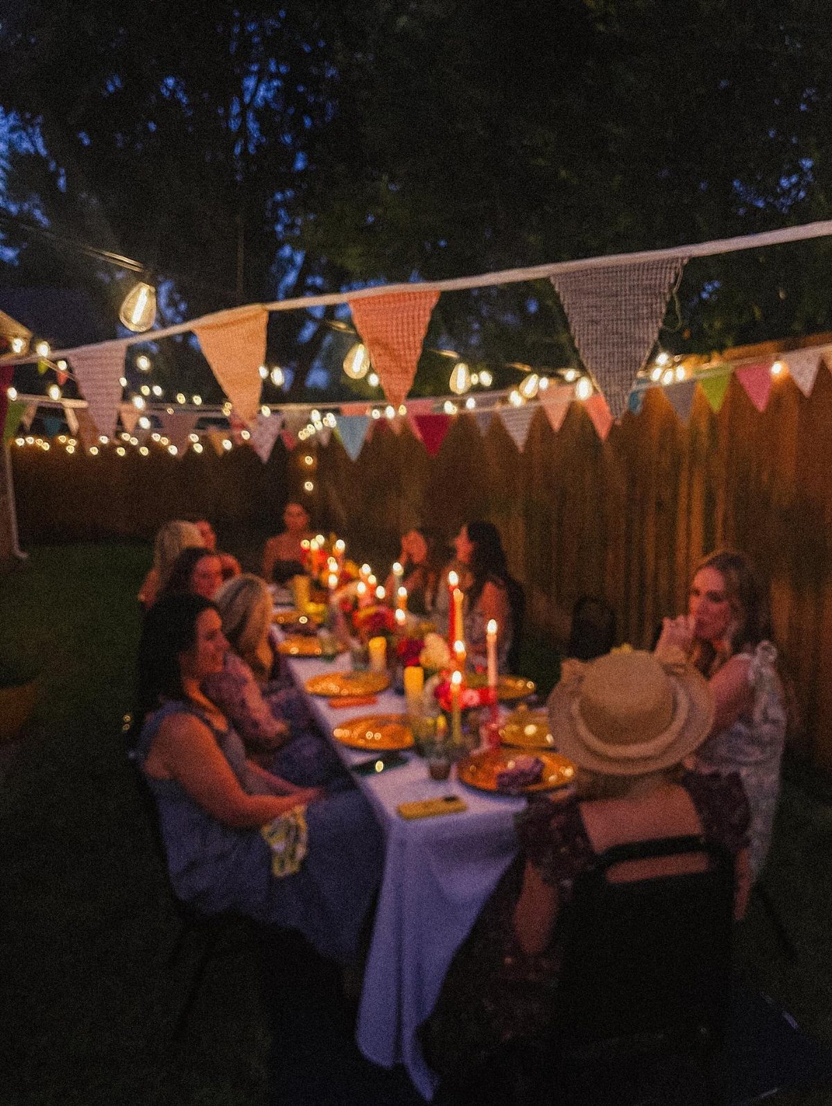 People dining at a long, candlelit table decorated with bunting in a backyard at night.