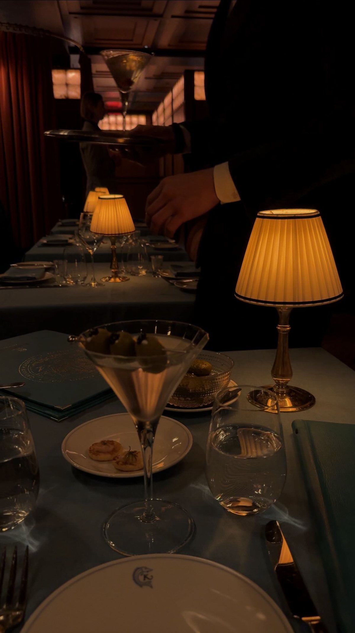 A formal dinner table set with small lamps, glassware, and a server carrying a martini.