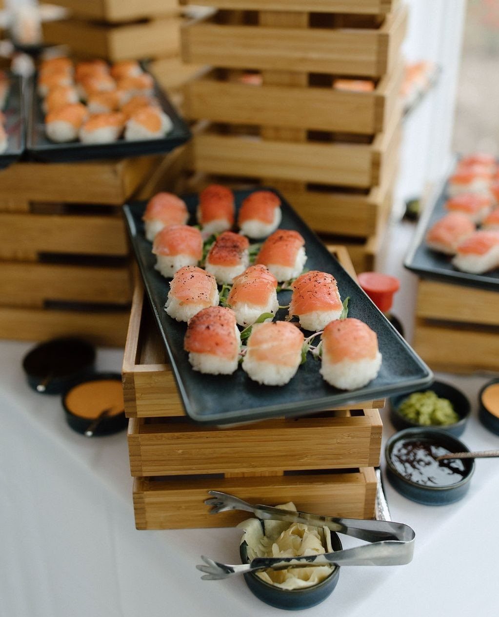Salmon nigiri sushi arranged on black plates stacked atop wooden crates on a white table.