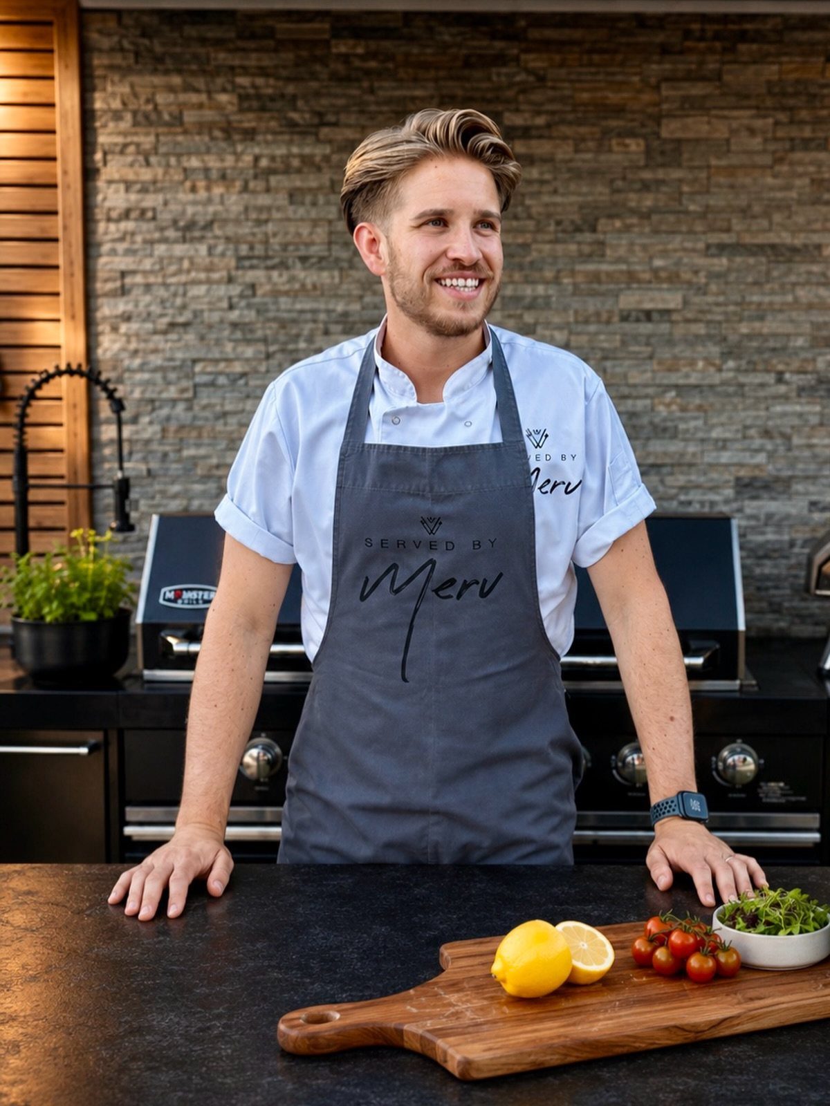 A smiling chef in a grey apron standing behind a kitchen counter with fresh ingredients.