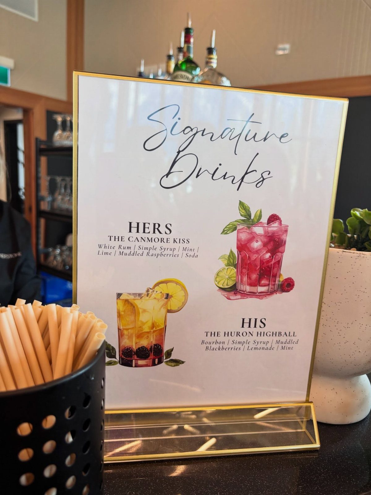 A white signature drinks menu sign with cocktail illustrations on a dark countertop.