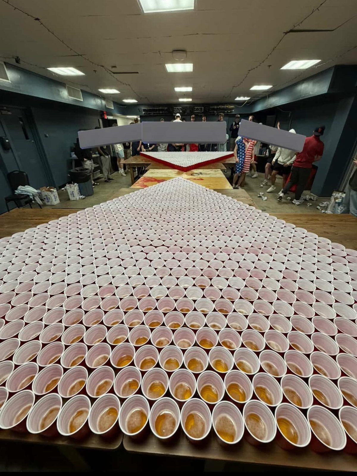 Massive triangular formation of white plastic cups filled with beer on a wooden floor.
