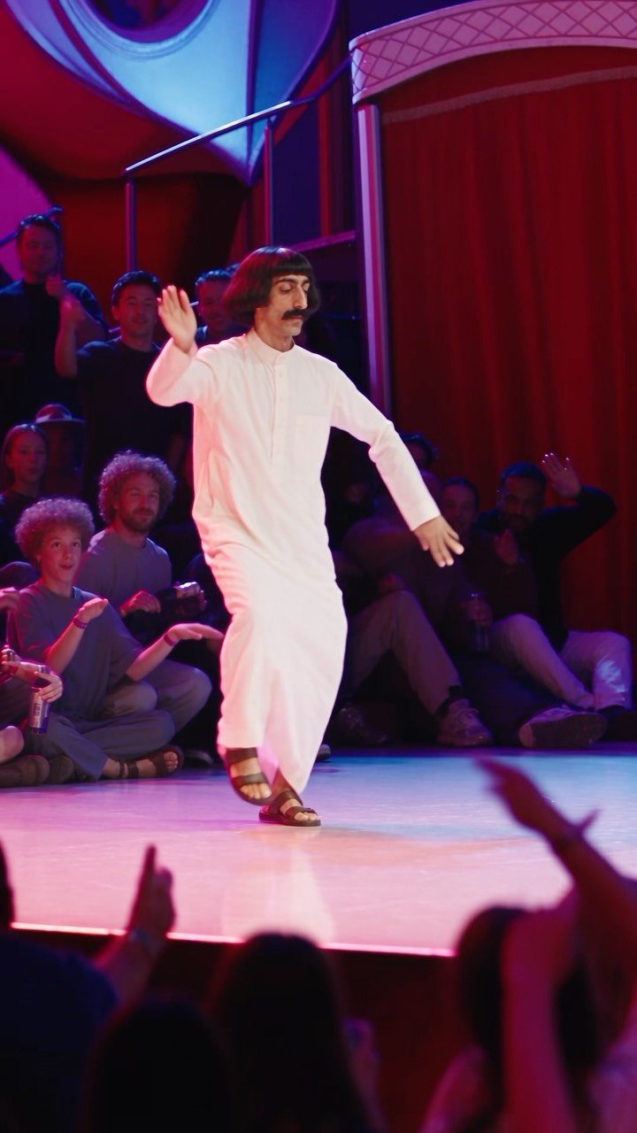 A man in white clothing dancing energetically on a brightly lit stage before an audience.