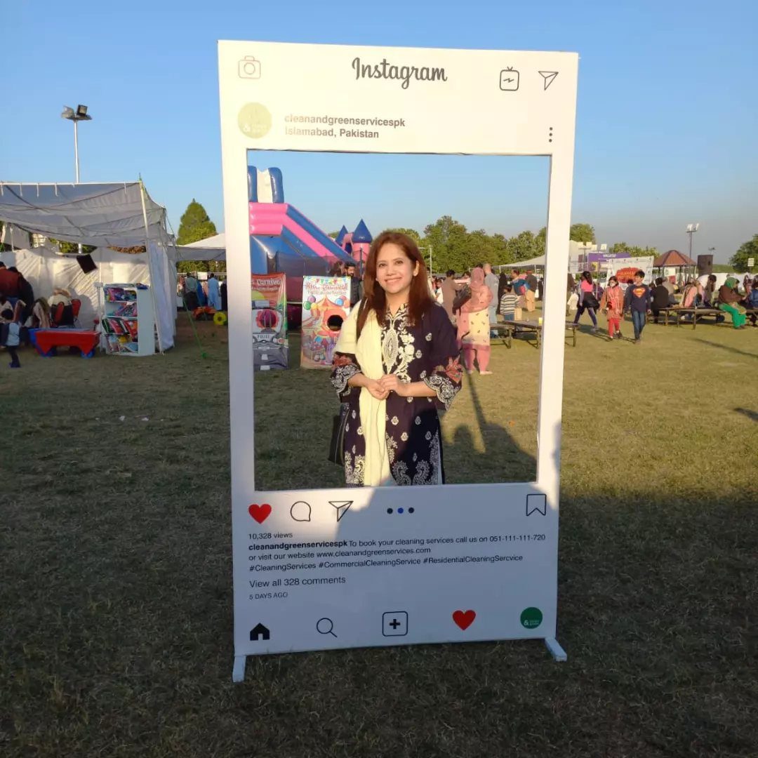 Woman posing inside a large white Instagram-themed photo booth frame on a grassy field.