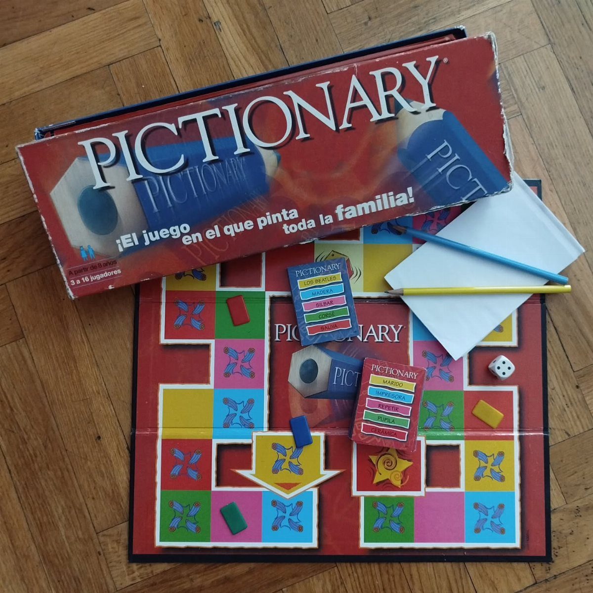 Pictionary board game components including the box, board, cards, and pencils on a wooden floor.