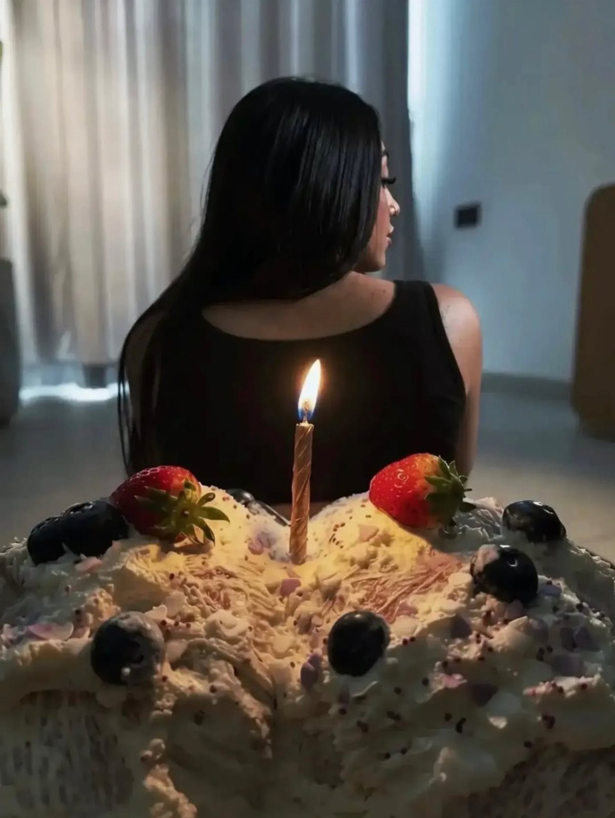 A decorated cream cake with strawberries and a lit candle in front of a woman.