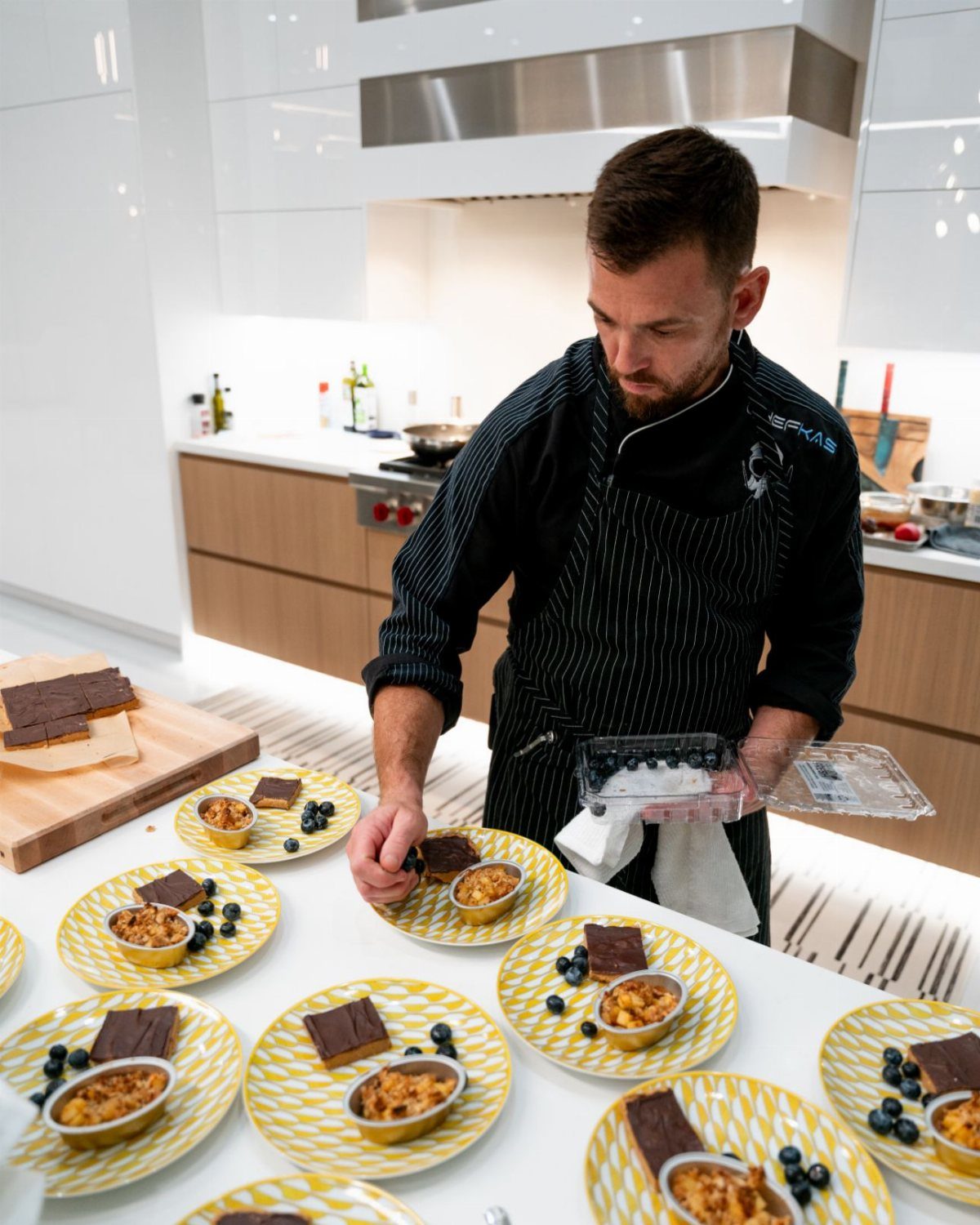 A professional chef plating gourmet chocolate desserts and blueberries on yellow patterned plates in a kitchen.