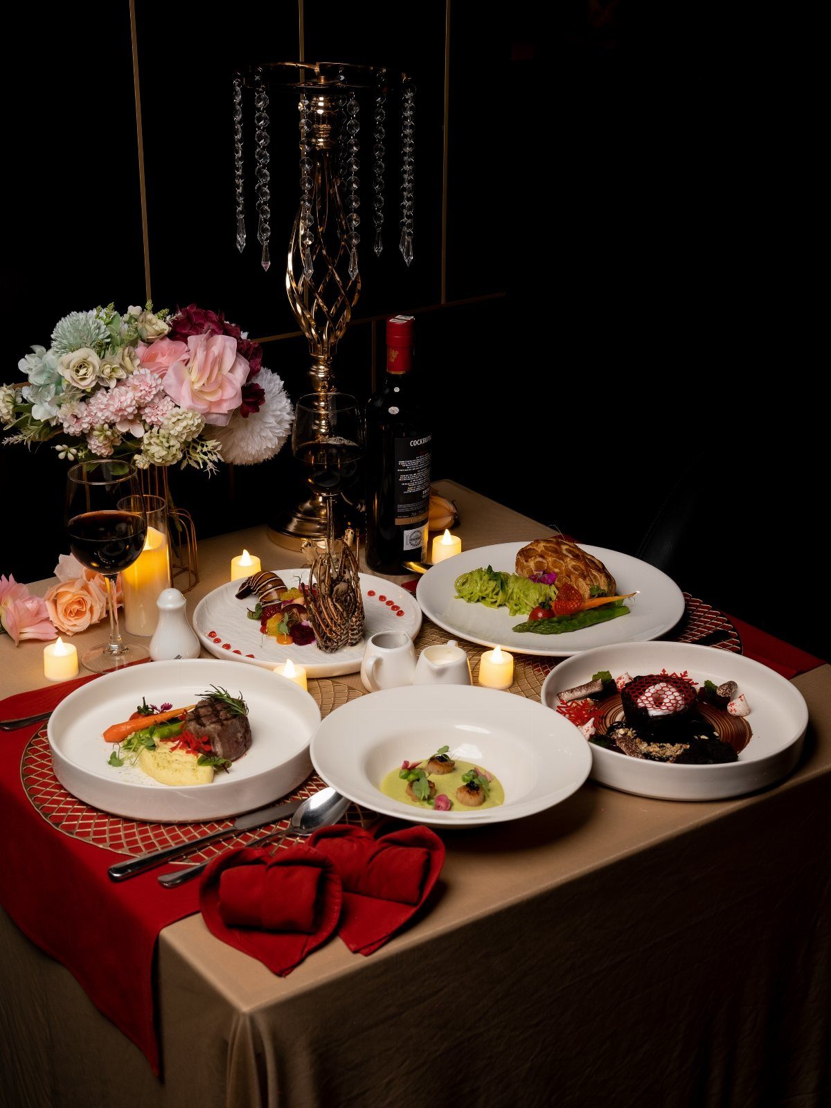 A gourmet dinner table set with multiple plates, wine, flowers, and lit candles.