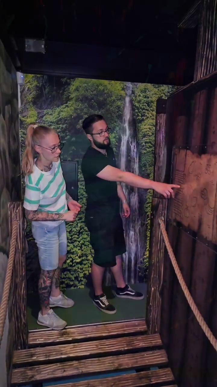 A man pointing at a wooden panel inside a jungle-themed escape room.