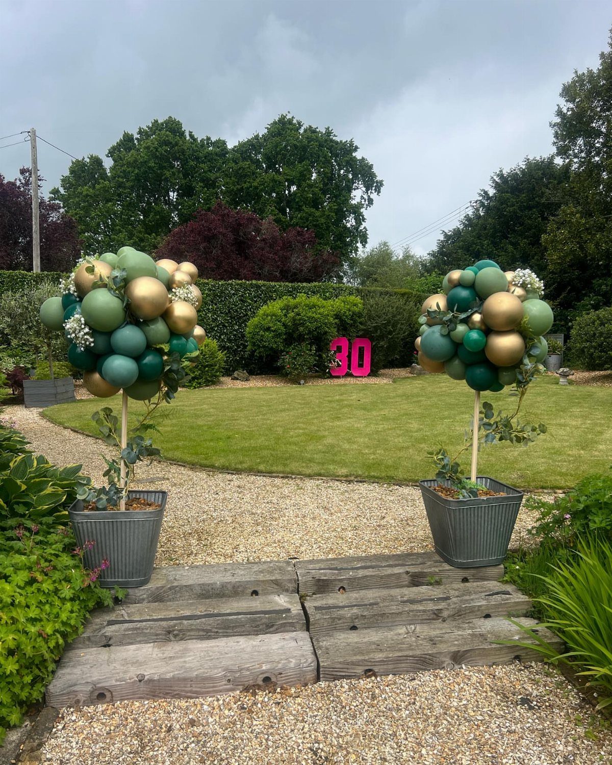 Green and gold balloon arrangements stand on a gravel path leading to a lawn with pink 30 decorations.