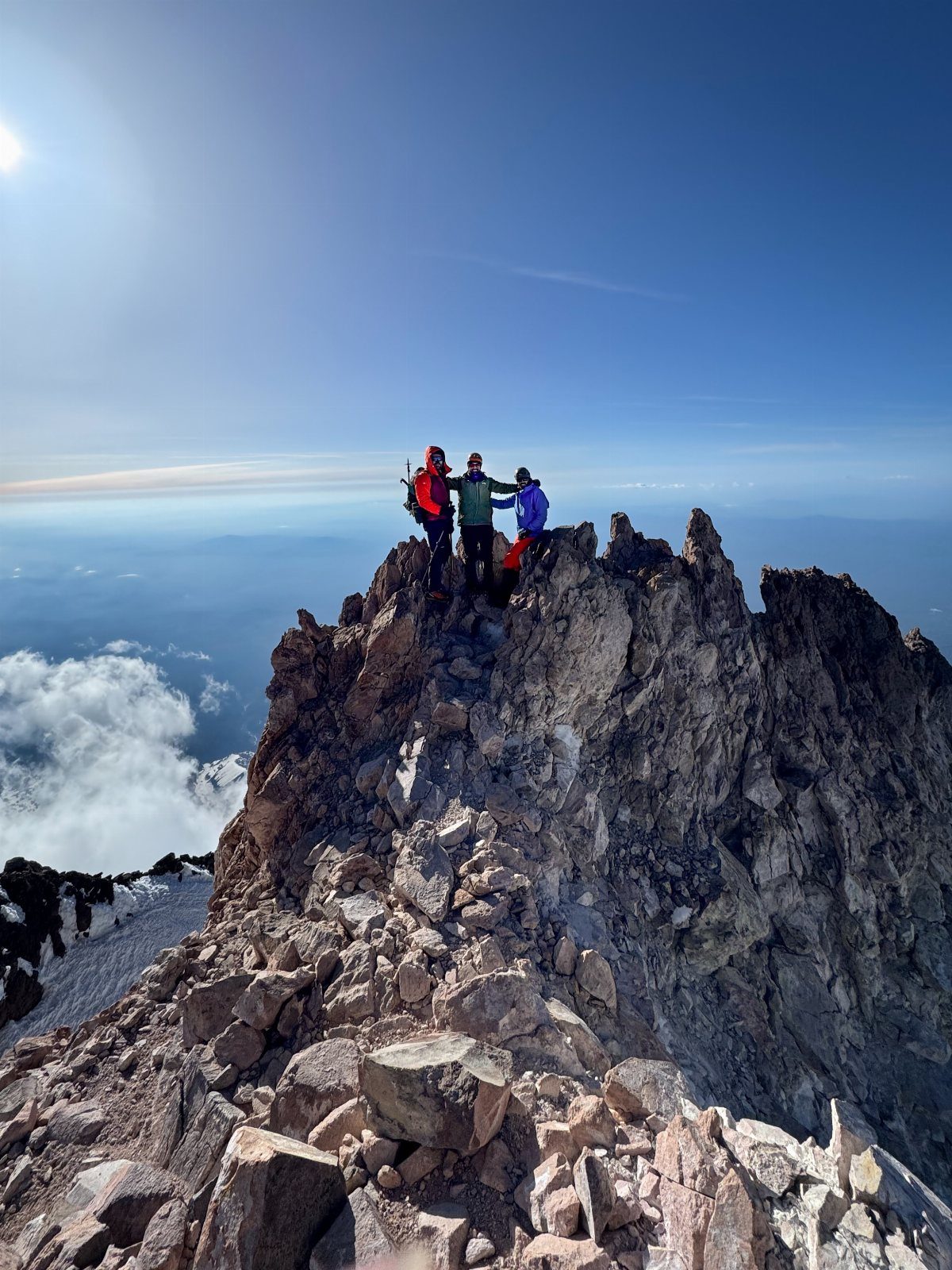 Three hikers standing on a rocky mountain summit above a sea of clouds.