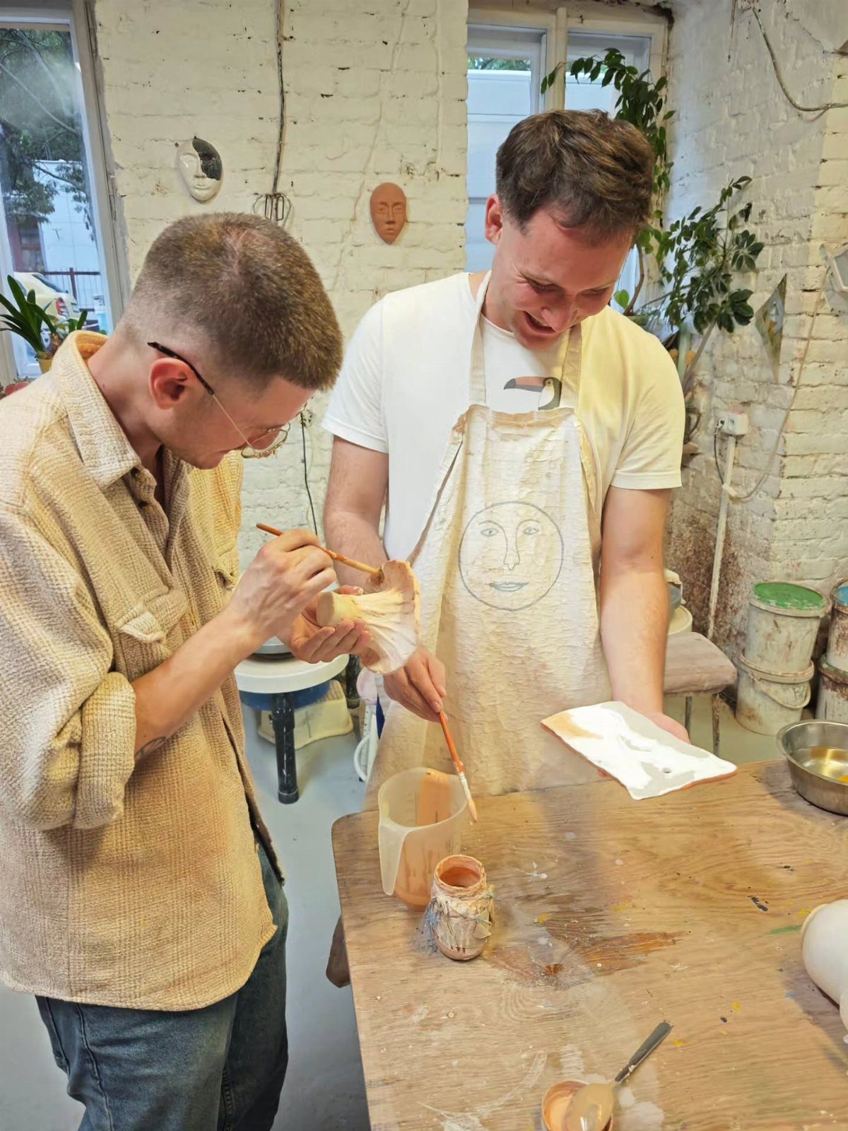 Two men focused on painting small ceramic objects at a wooden pottery workshop table.