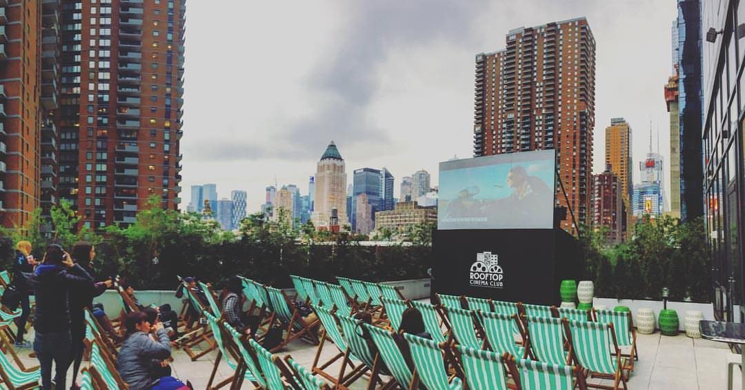 Striped deck chairs arranged on a rooftop facing a large movie screen against a city skyline.