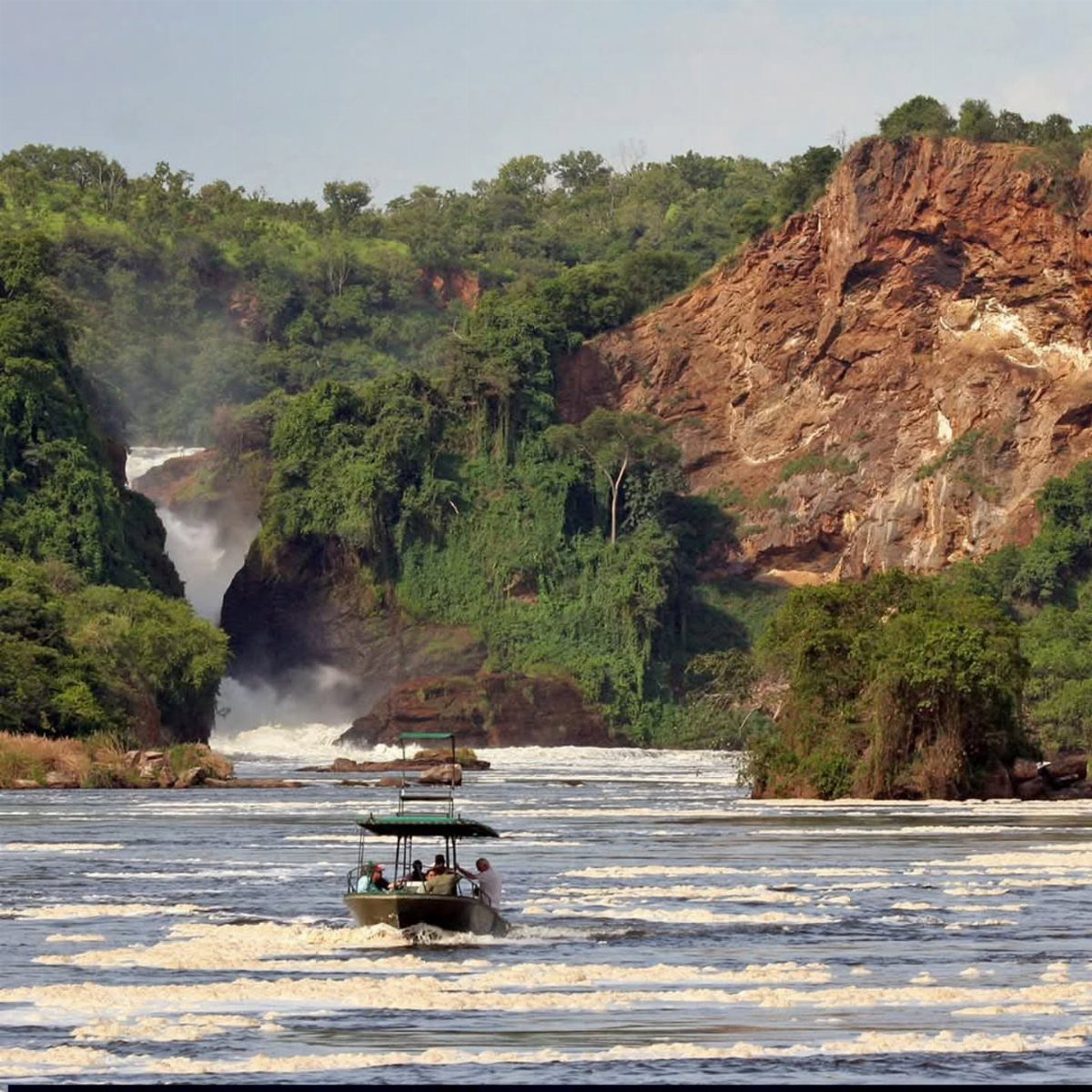 A small boat carrying people navigates through river rapids near a large waterfall.