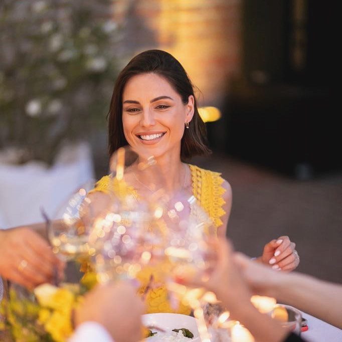 A smiling woman in a yellow top toasts with friends using wine glasses outdoors.