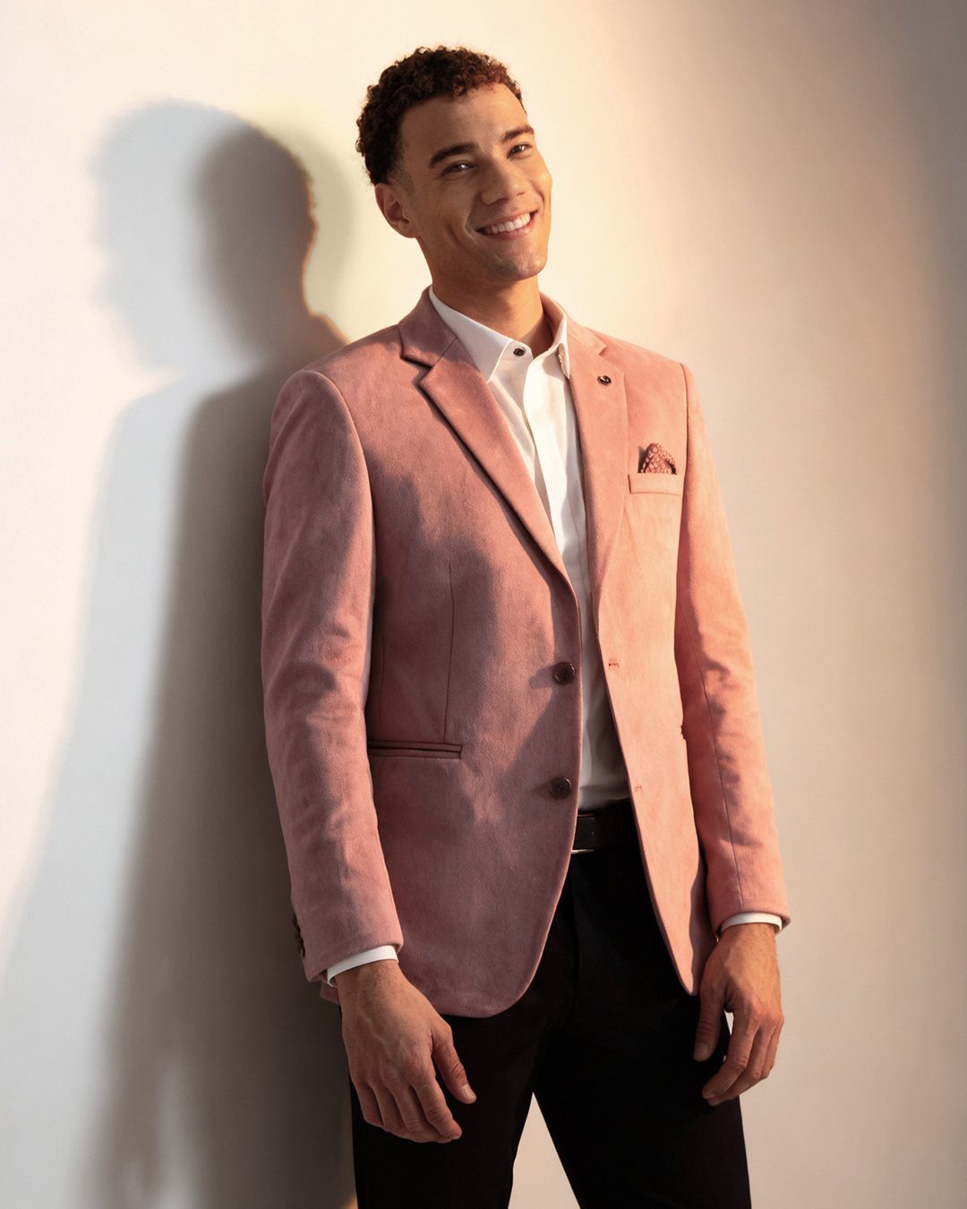 A smiling man posing in a light pink textured blazer and white shirt.