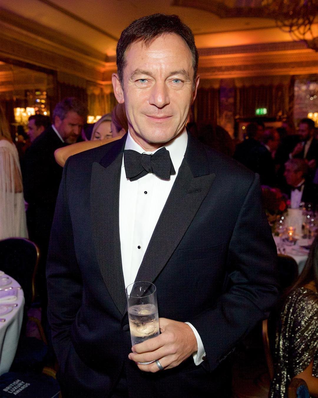 A man wearing a formal black tuxedo and bow tie holding a glass of water.