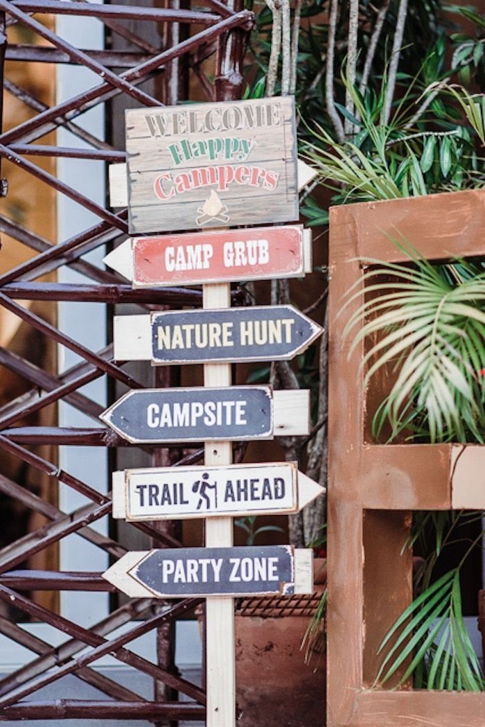 Wooden directional signpost with camping-themed text standing amidst green plants and a dark lattice.