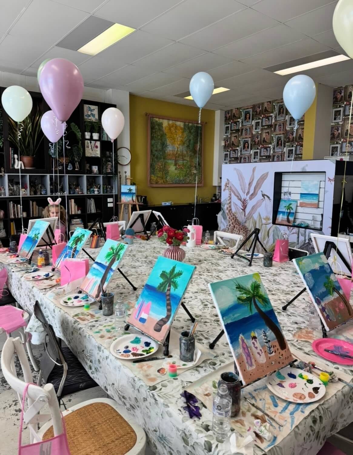 Small tropical beach paintings on easels arranged on a long table with art supplies.