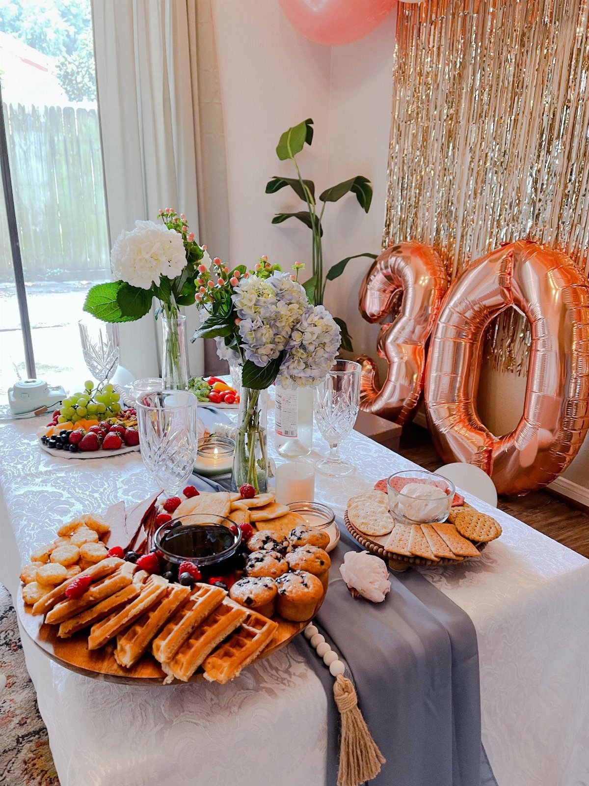 A festive brunch table set with waffles, pastries, fruit, and rose gold number thirty balloons.