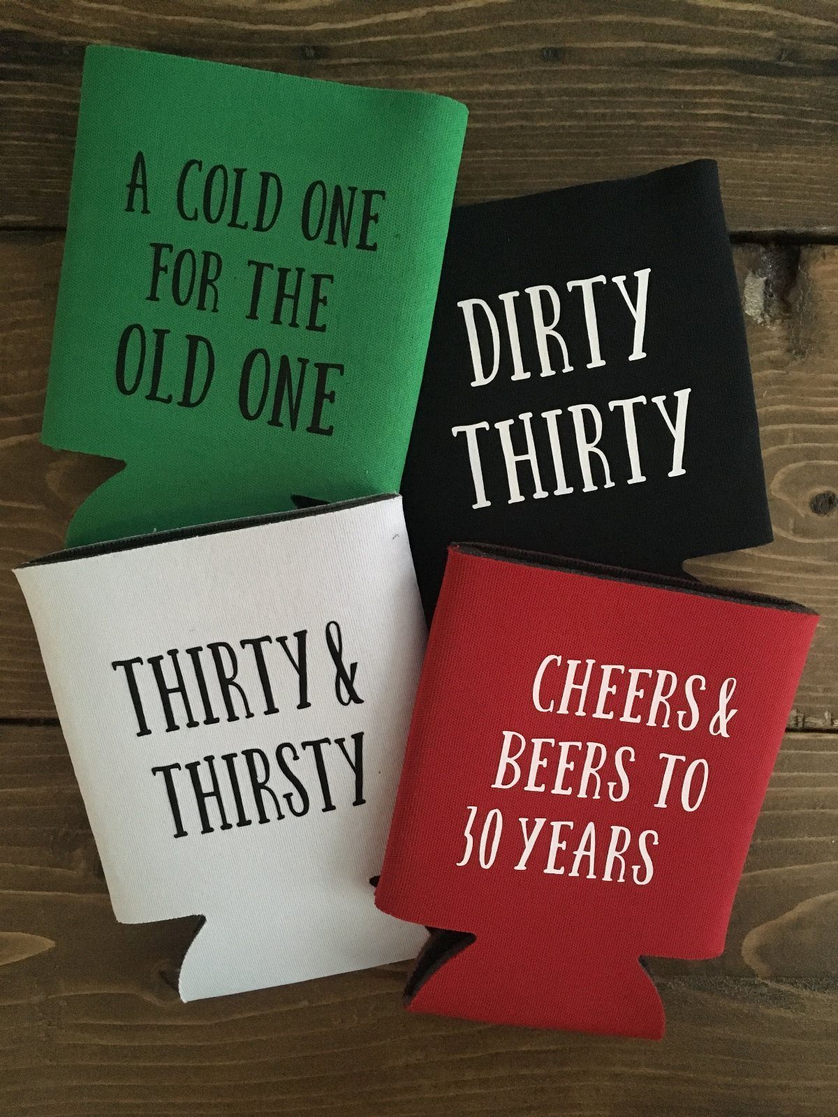 Four colorful beverage can insulators with 30th birthday slogans resting on a wooden table.