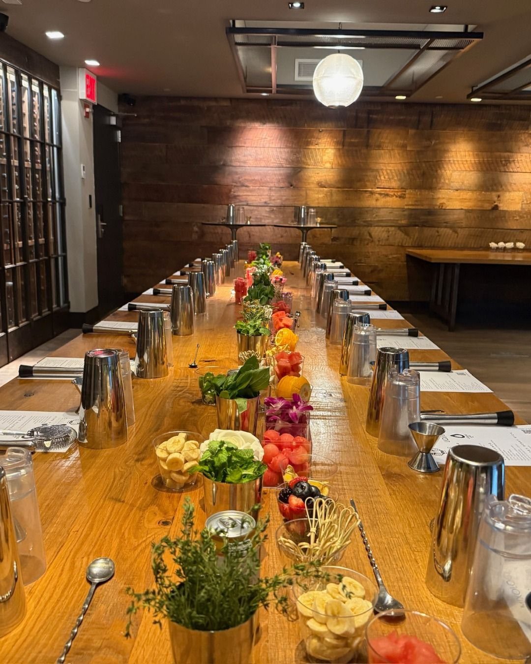 A long wooden table lined with silver cocktail shakers and bowls of fresh fruit and herb garnishes.