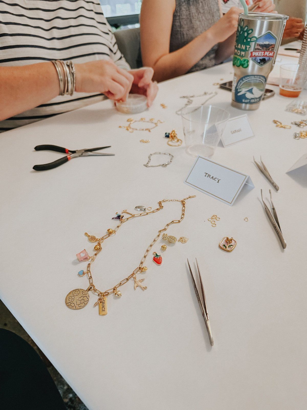 People assembling gold charm necklaces with pliers and tweezers on a white table.