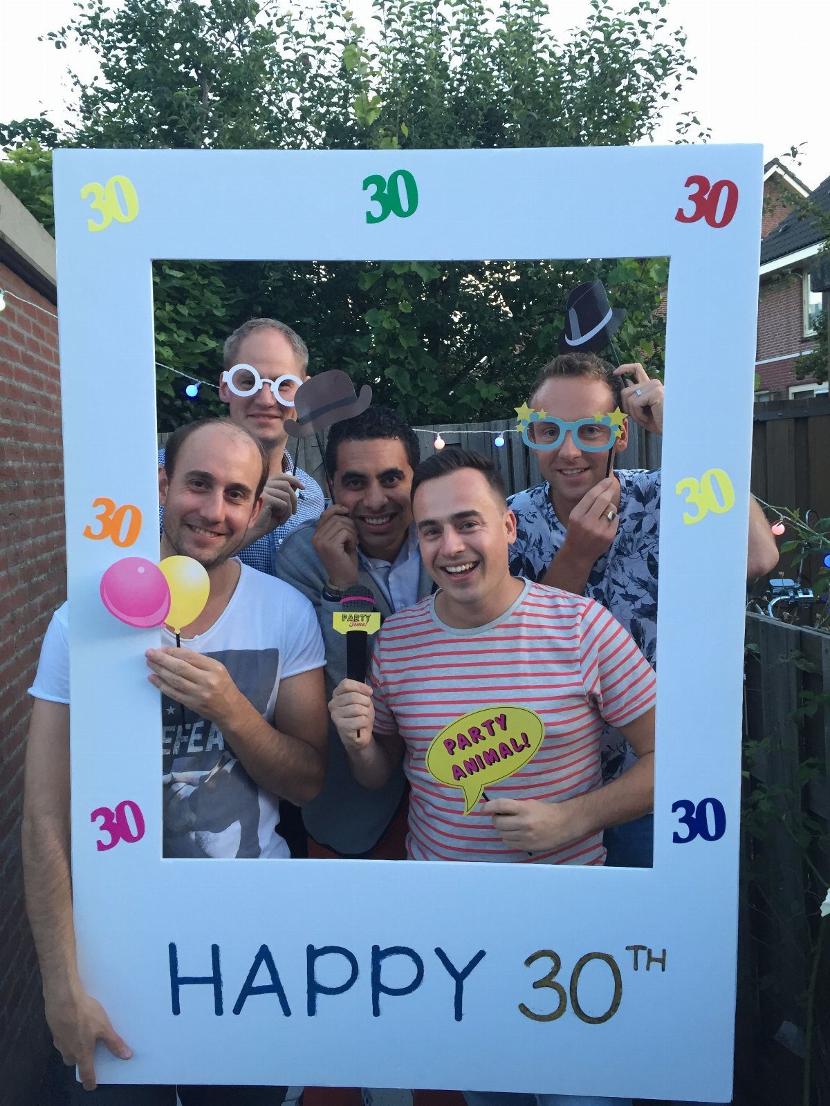 Five men smiling behind a large blue DIY photo booth frame decorated with happy 30th birthday text.